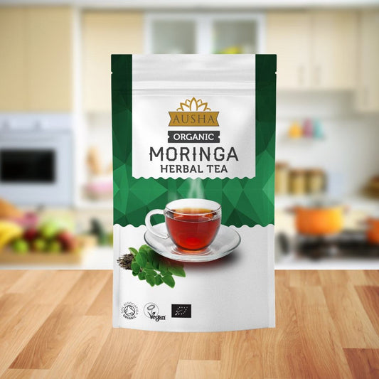 Organic Moringa Tea 100g packaging on a kitchen countertop, featuring a cup of herbal tea with fresh moringa leaves.
